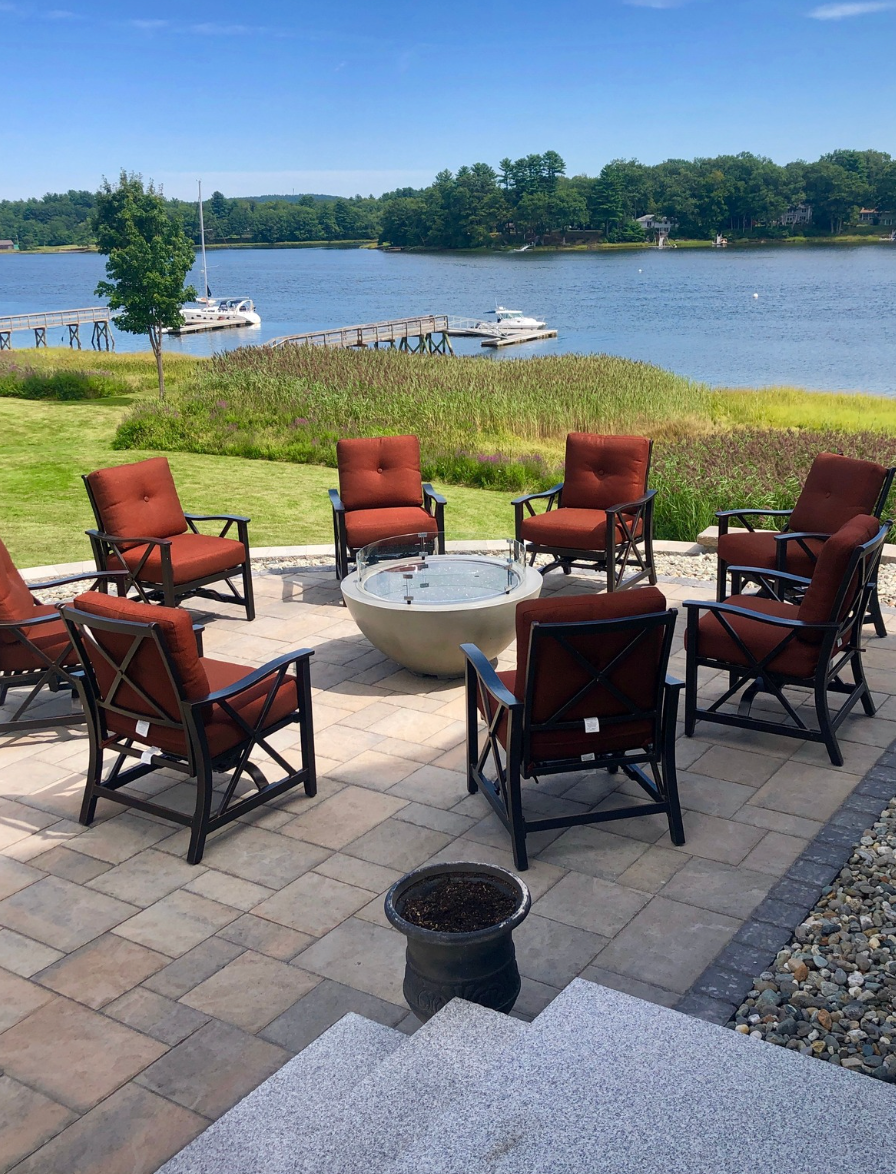 Patio chairs around a fire pit overlooking a calm lake on a sunny day.