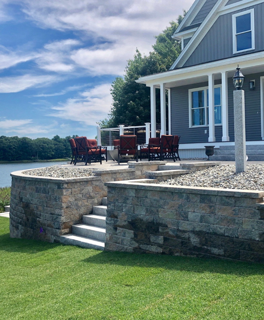 Stone patio beside a gray lakeside house with red chairs, white umbrella, and lawn steps.