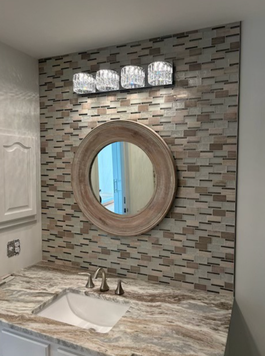 Bathroom vanity with mosaic tile wall, round mirror, and three-light fixture above a marble countertop.