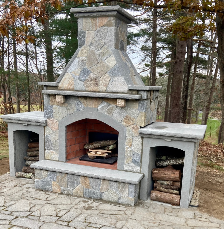 Stone outdoor fireplace with wood-burning firebox and side storage niches in a wooded yard.