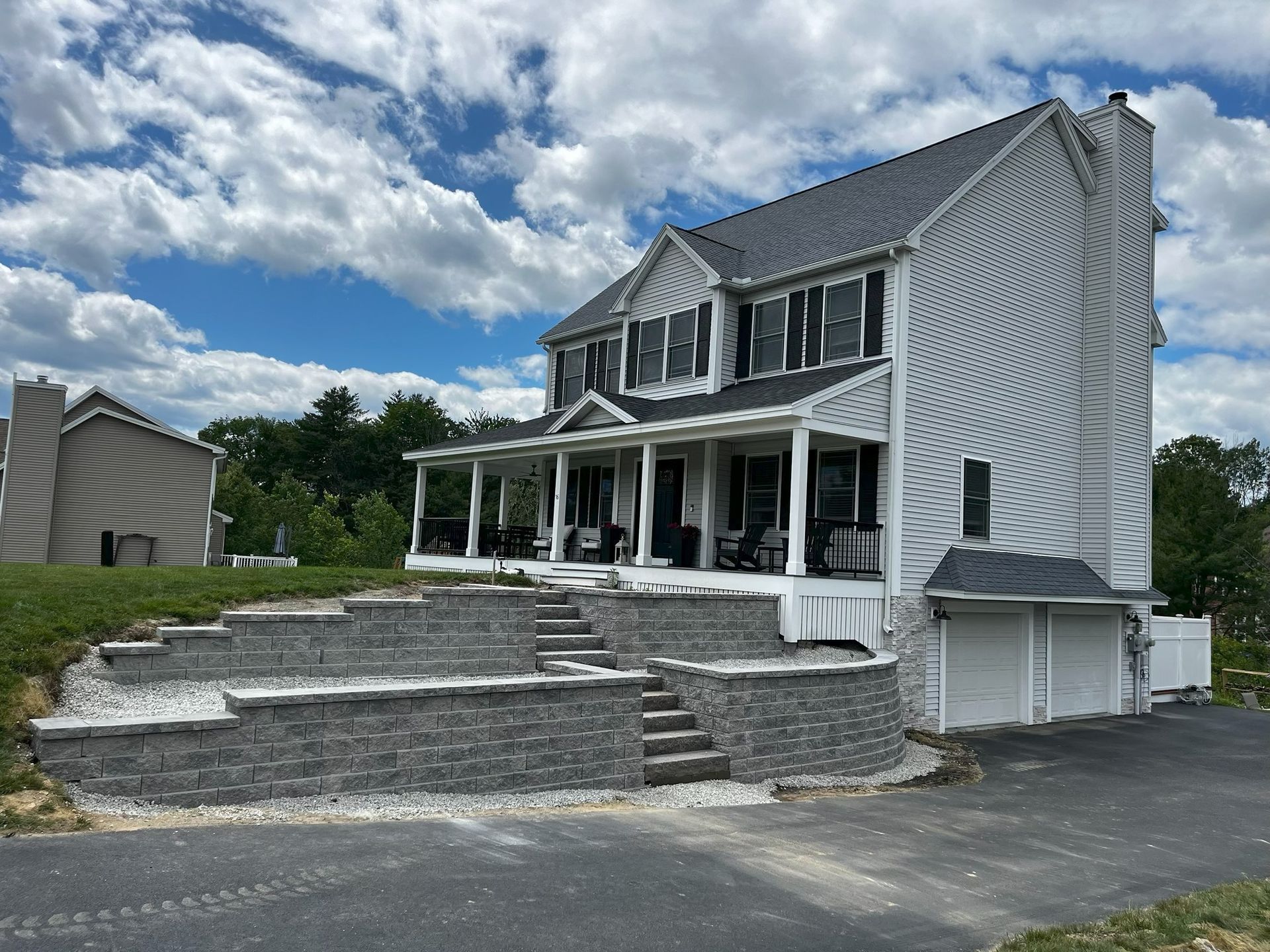 Two-story gray house with front porch, stone retaining wall, and cloudy sky.