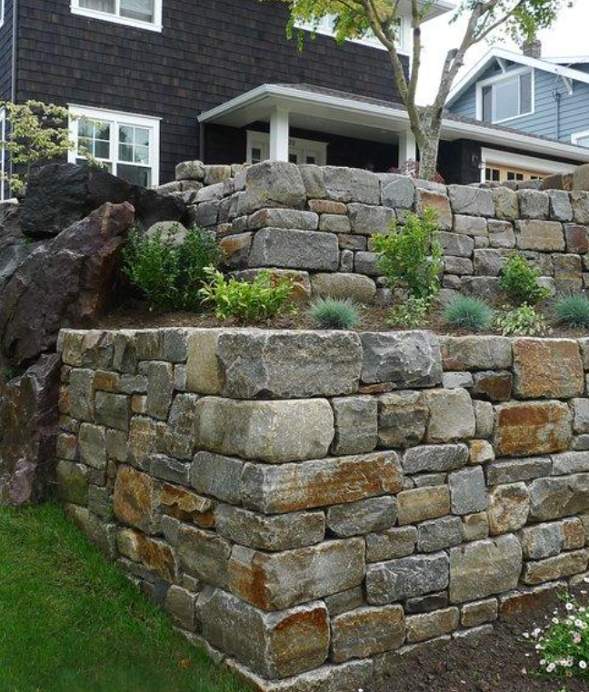 Stone retaining wall terraced with shrubs in front of a dark-sided house