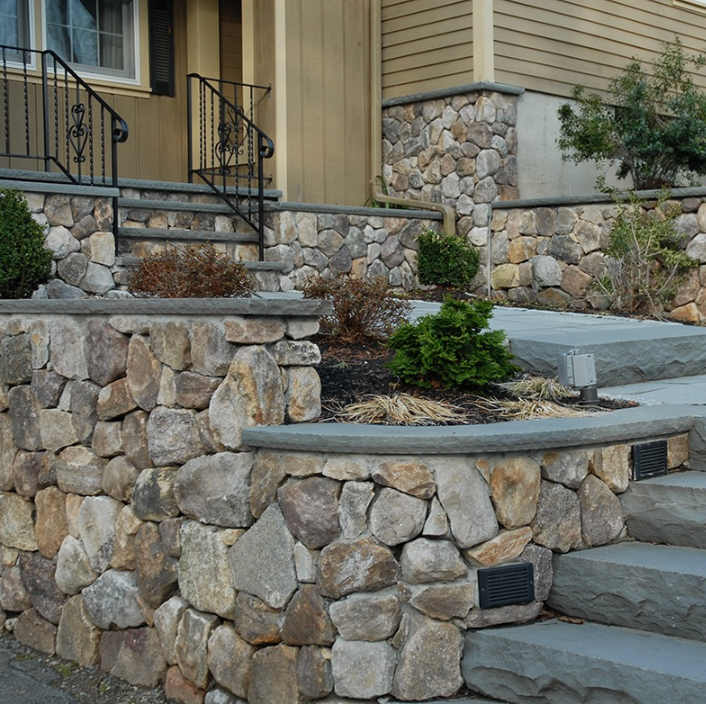 Stone retaining walls and concrete steps leading to a house entrance with black railings and shrubs.