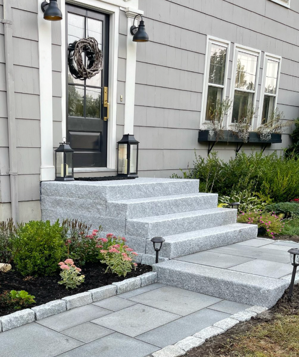 Front entrance with gray stone steps, paver walkway, lanterns, and flower beds beside a house