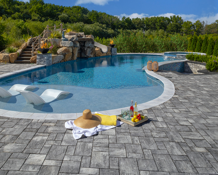 Backyard pool with stone patio, waterfall feature, white lounge chairs, and a straw hat on a towel by snacks