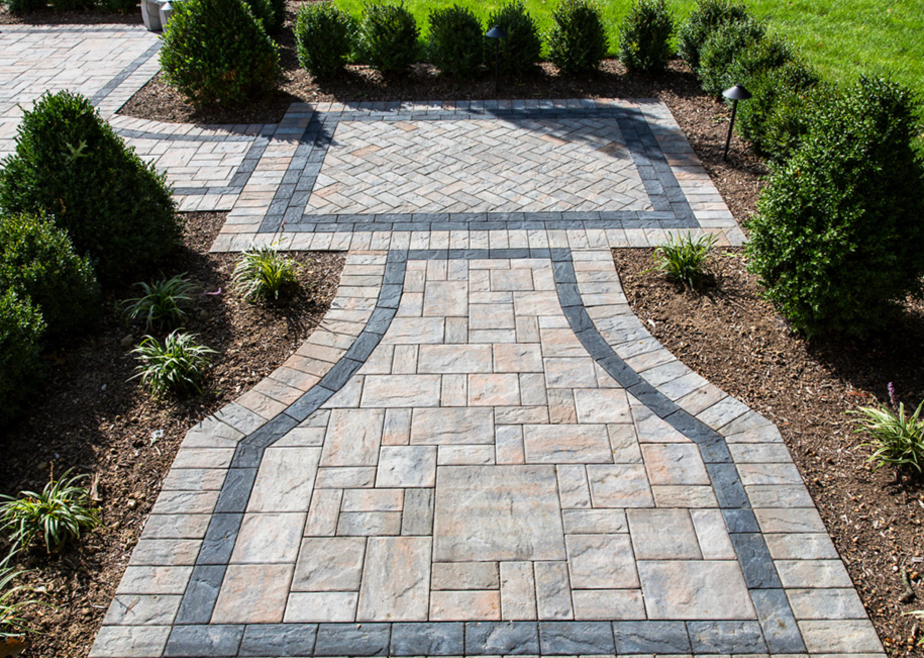 Curved stone walkway leading to a patio, bordered by gray pavers and trimmed shrubs in a landscaped yard