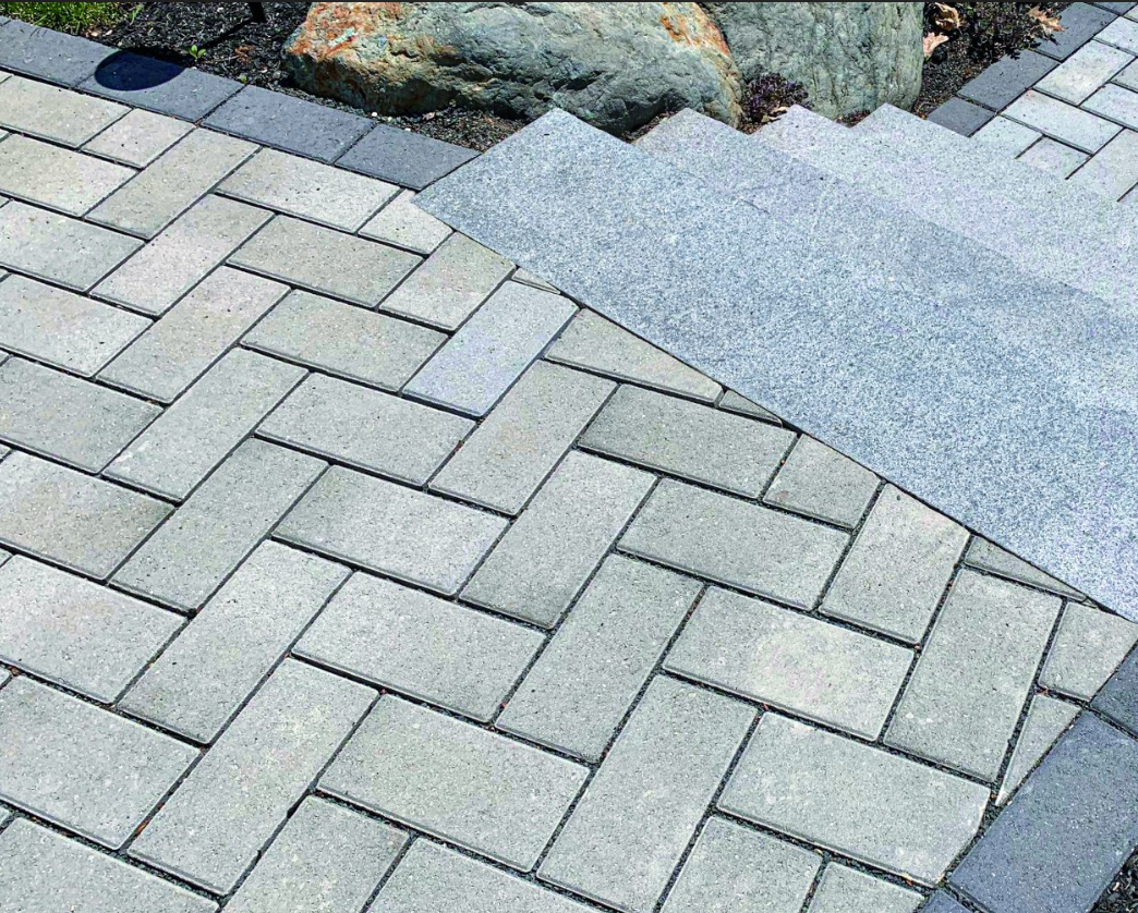 Gray interlocking pavers beside a rough concrete path and a small planted border
