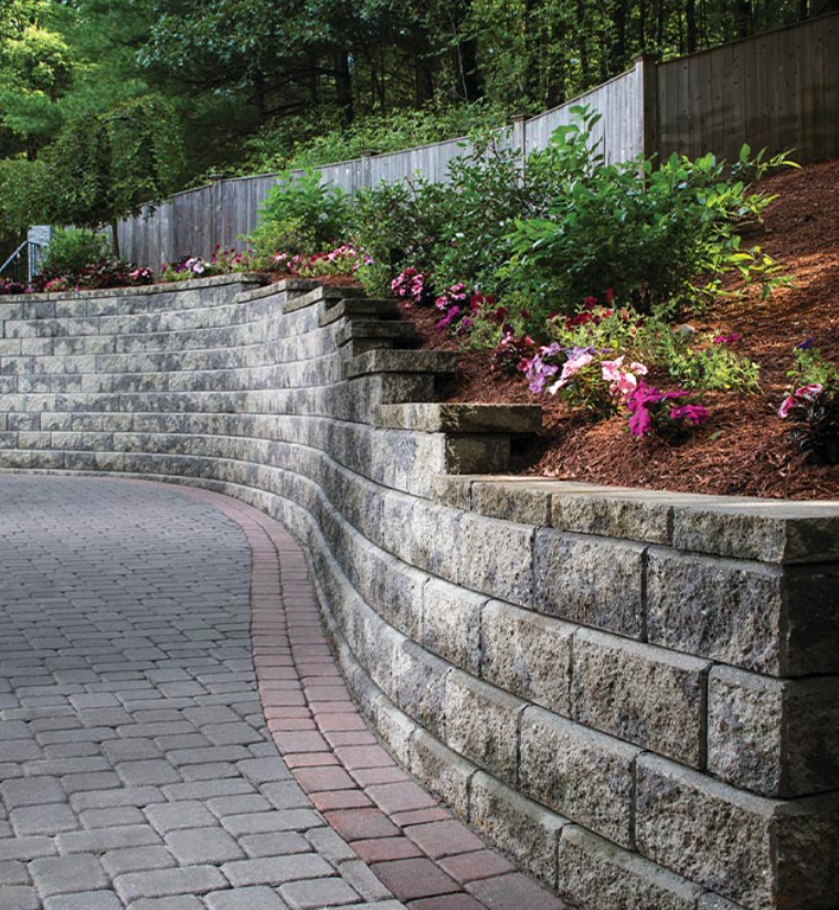 Curved stone retaining wall beside a paved path, with pink flowers and greenery on a hillside.