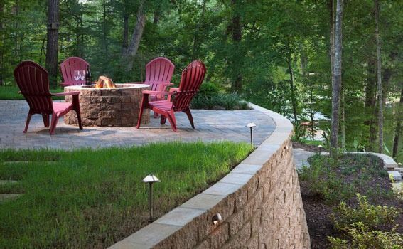 Stone patio with red chairs and a fire pit beside a wooded creekside lawn