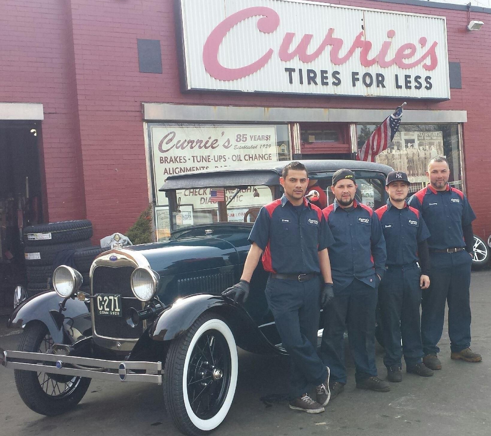 a group of men standing in front of a car in front of a store called currie 's tires for less