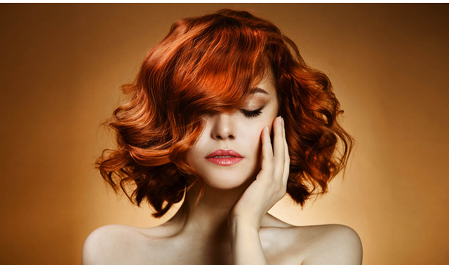 Woman with reddish-orange wavy hair, hand near face, eyes closed, on a warm brown background.
