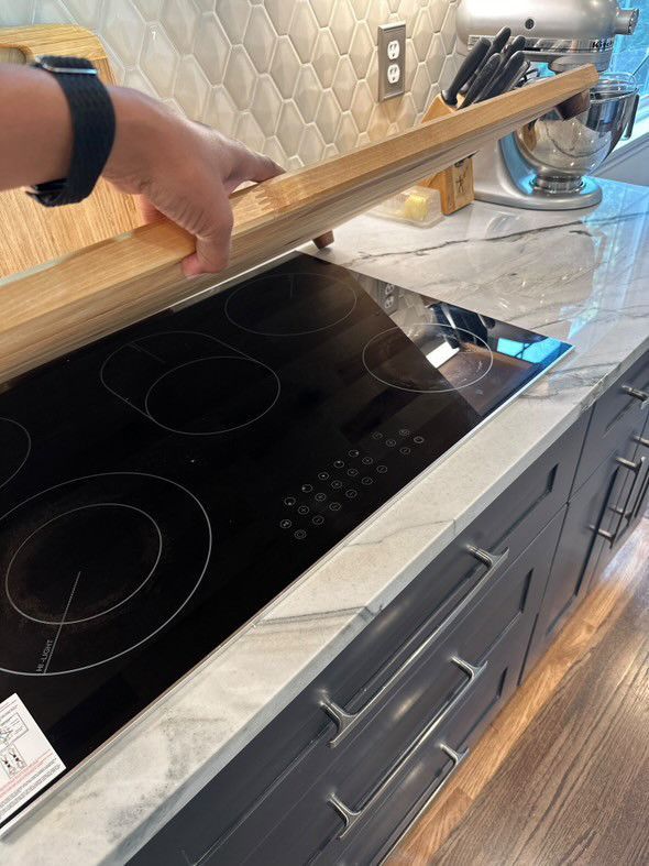 A person is holding a wooden cutting board over a stove in a kitchen.