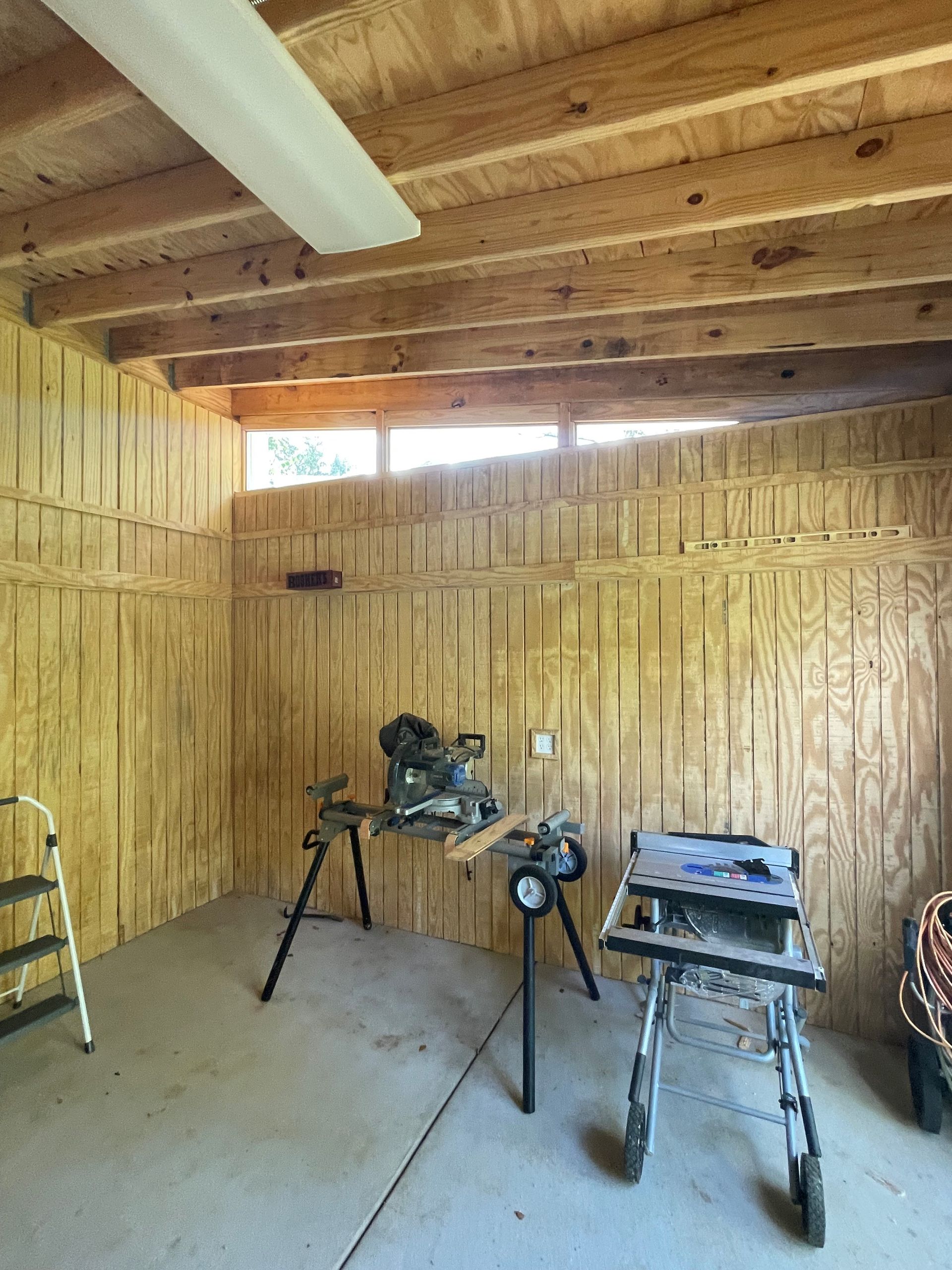 A wooden garage with a table saw and a ladder.