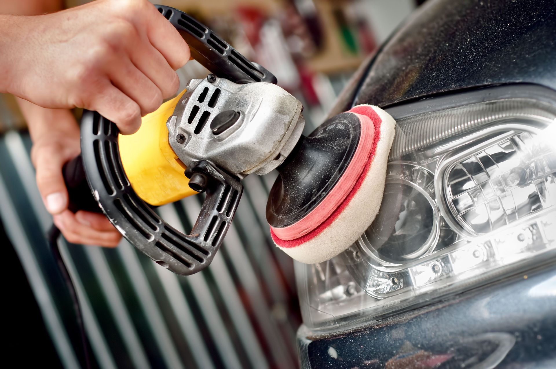 Car polishing a headlight with a buffer and red pad, close-up on the front of a dark vehicle