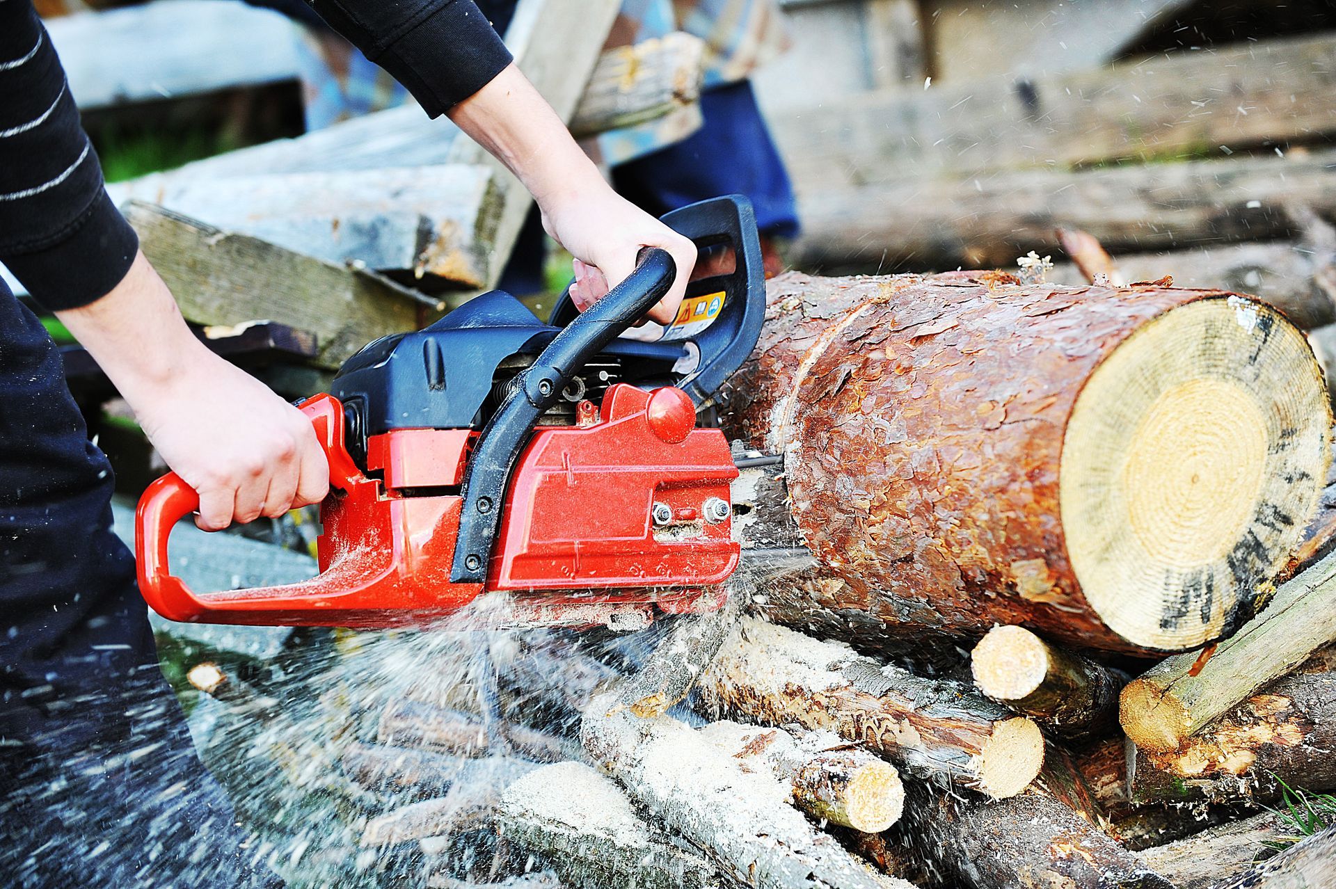 Hands using a red chainsaw to cut a large log, with wood chips flying on the ground.