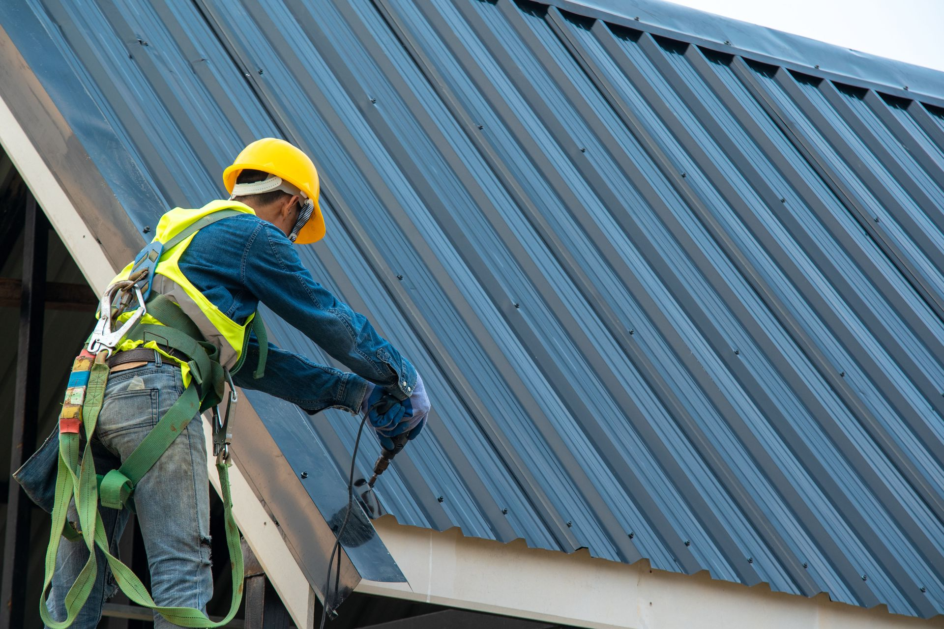 Worker in a yellow hard hat and safety harness cleaning a blue corrugated metal roof.