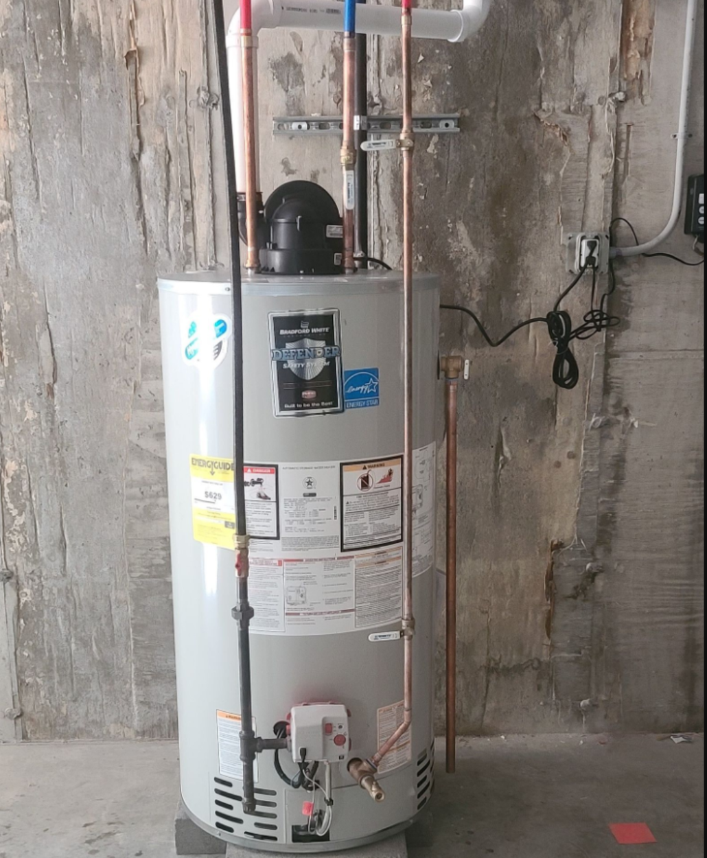 Water heater against a weathered concrete wall, with copper pipes and electrical components.