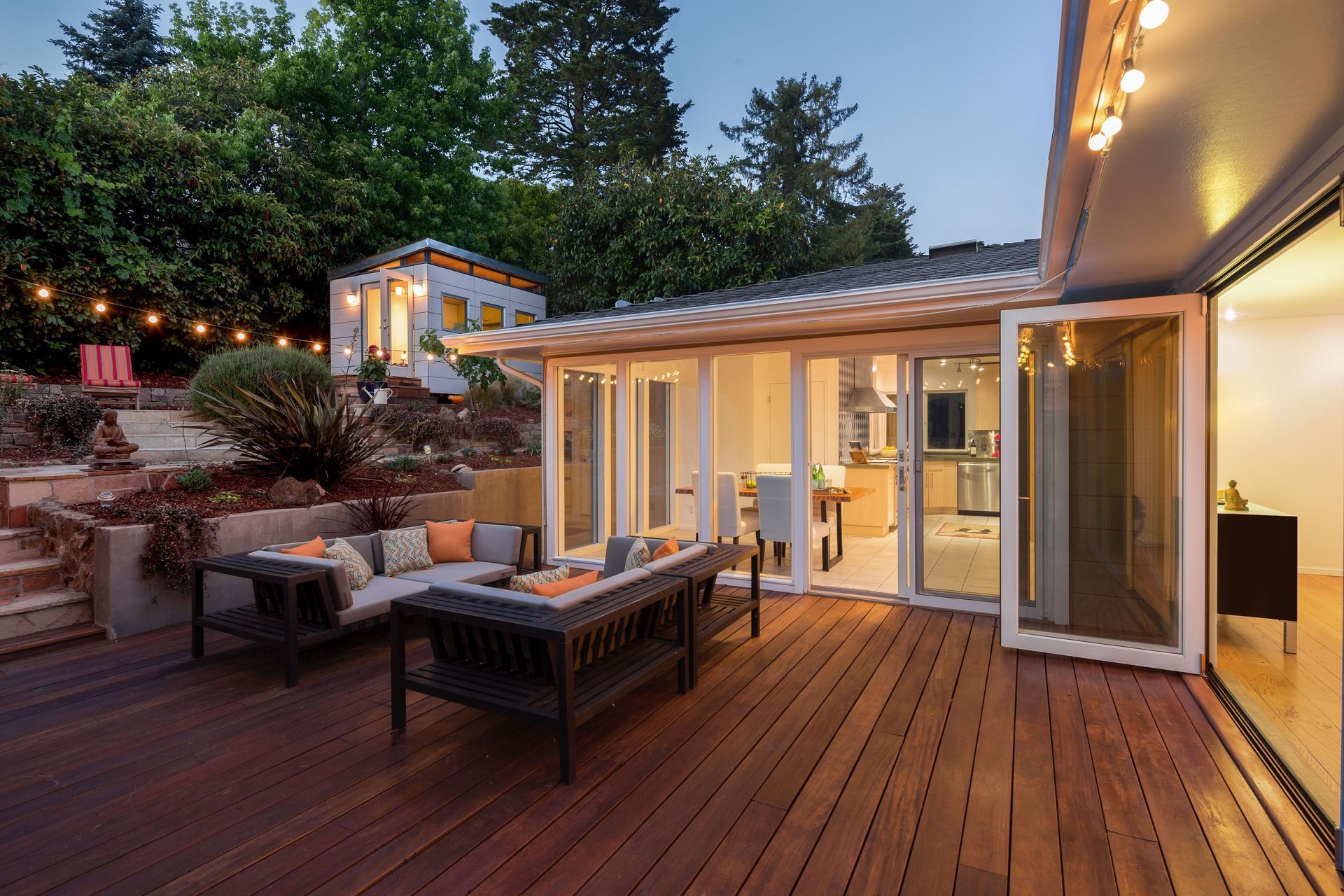 Wooden deck patio at dusk with outdoor seating, string lights, and open glass doors to a lit house