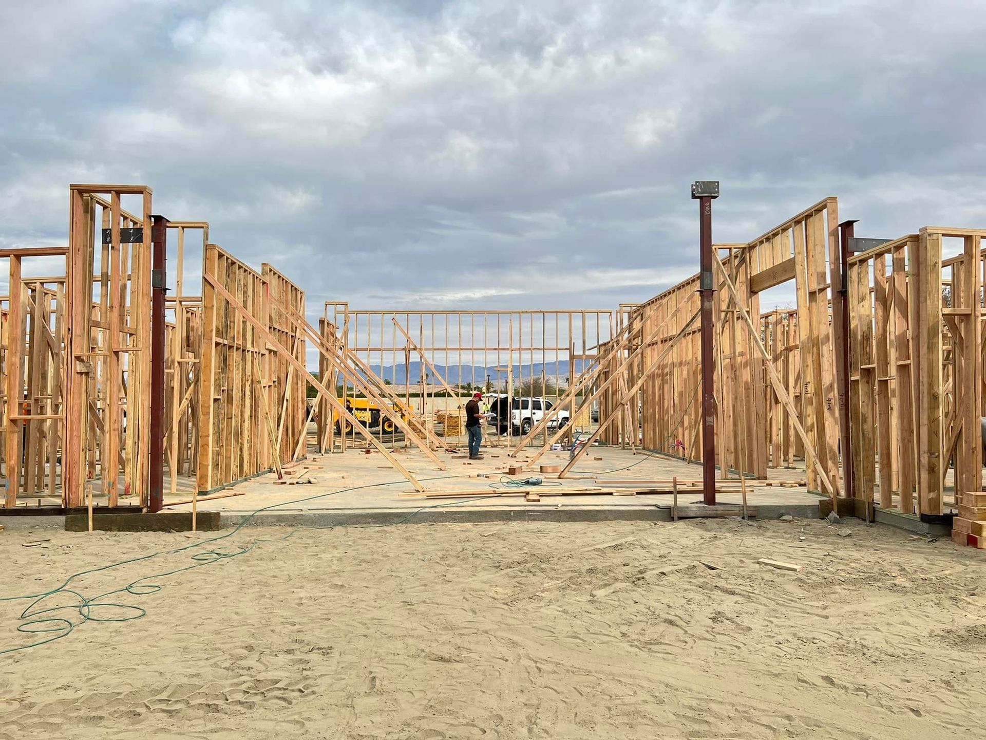 Wood-framed building under construction on a sandy lot under a cloudy sky