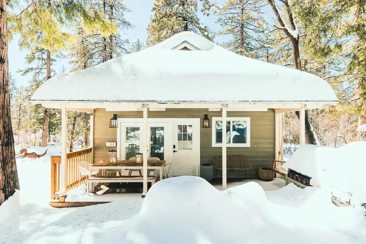 Snow-covered cabin porch with white roof, wooden railing, and pine trees in bright winter sunlight