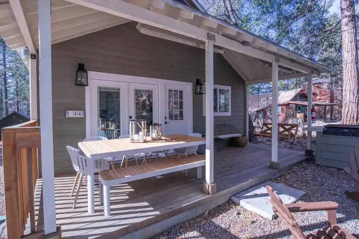 Covered porch with white table and chairs outside a gray house, surrounded by trees and gravel.