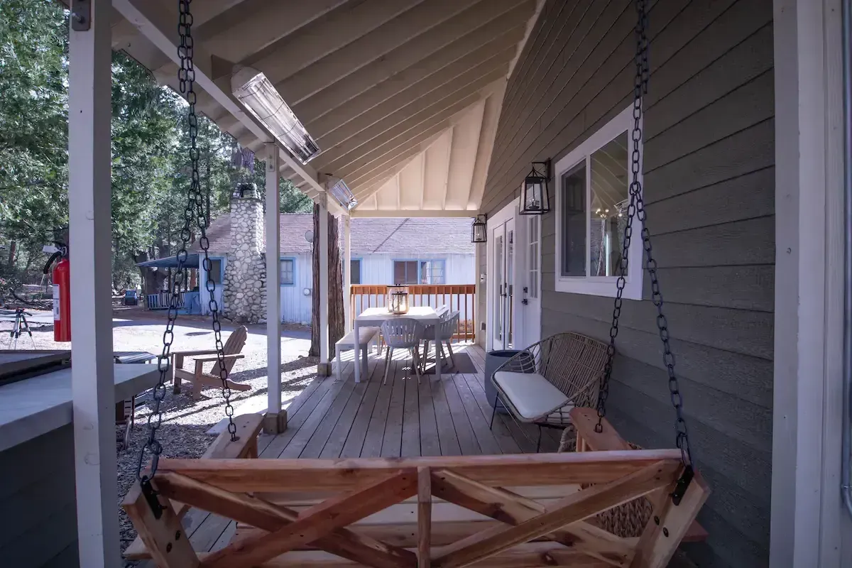 Covered porch with rocking chairs, a hanging swing, and a view of trees and a house across the yard.