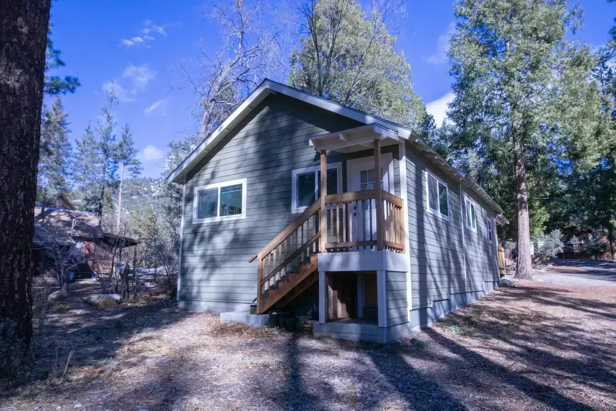 Small gray cabin with a wooden porch in a wooded area under a blue sky