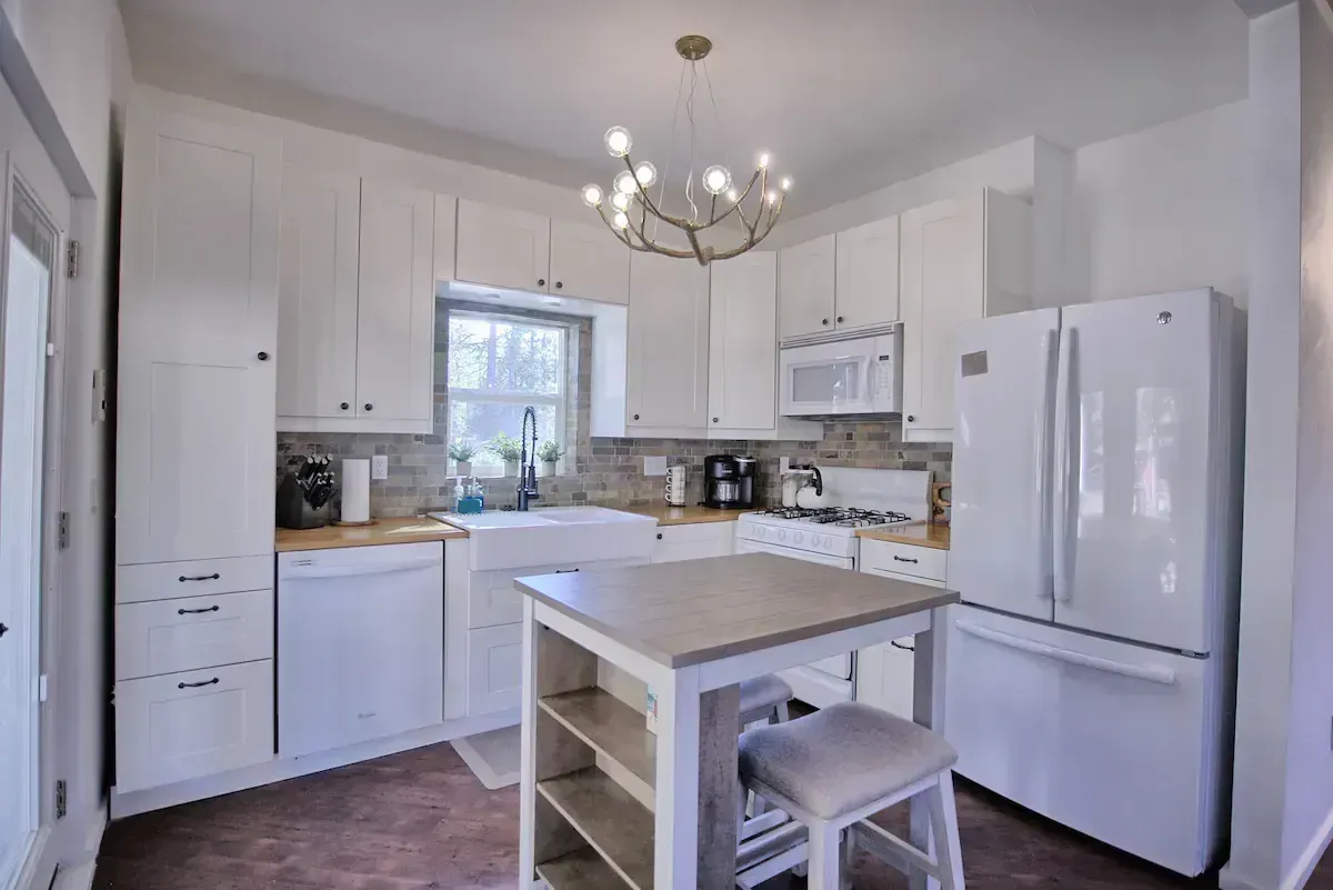 Bright white kitchen with island, chandelier, refrigerator, and window above the sink