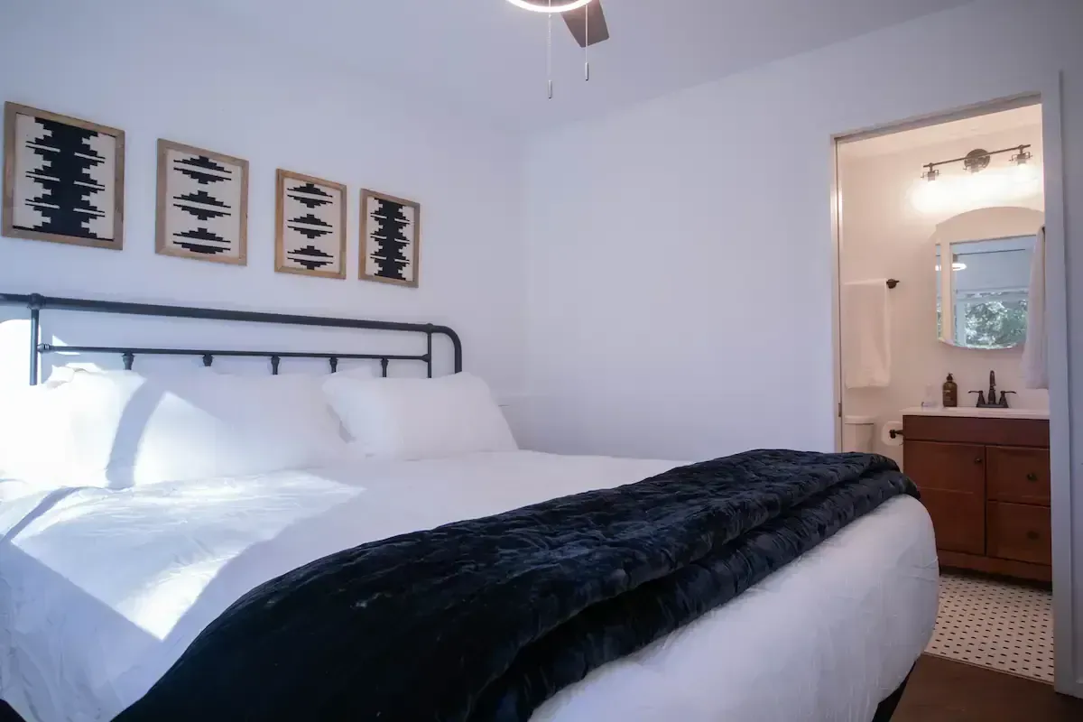 Bright bedroom with white bedding, dark throw blanket, wall art, and an open doorway to a bathroom.