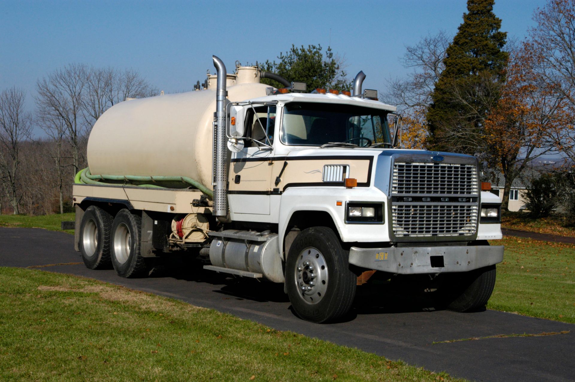 A beige and white tanker truck parked on an asphalt path next to a grass lawn under a clear sky.