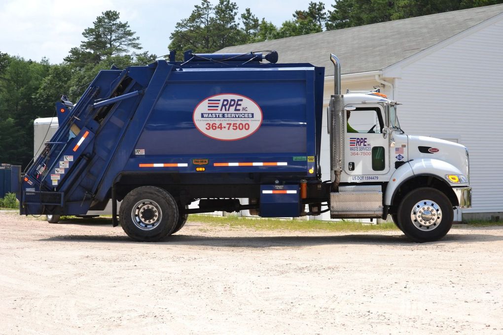 A blue and white RPE trash truck parked on a gravel lot near a building.
