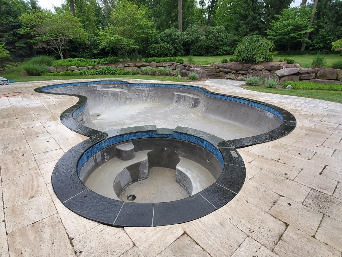 Empty kidney-shaped pool with a circular spa, surrounded by stone patio. Green trees in background.