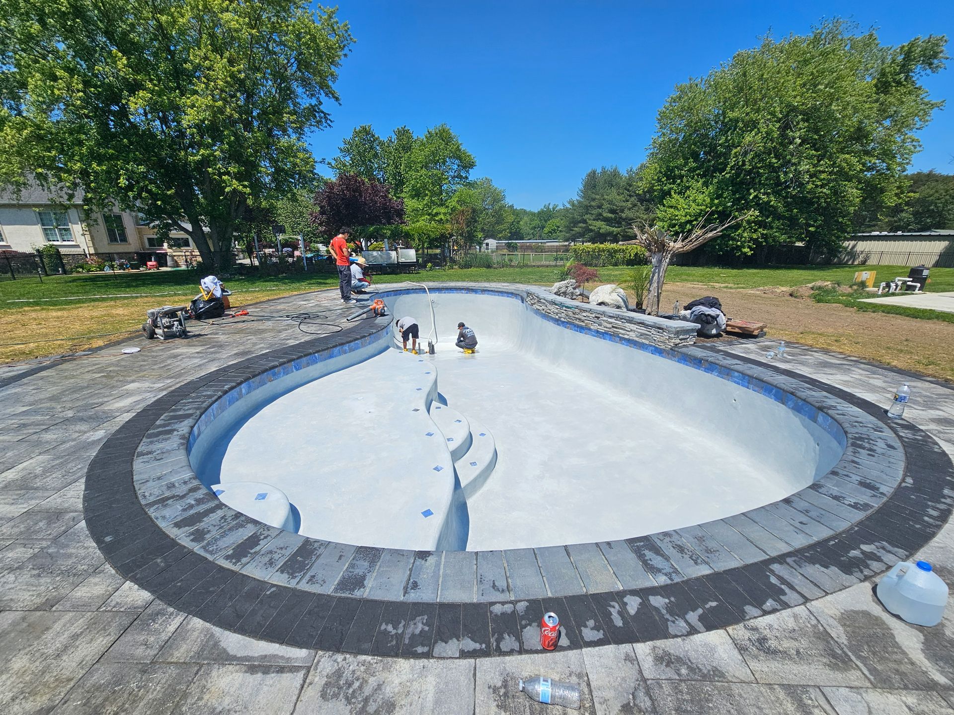 Workers installing tiles around a kidney-shaped pool. Blue sky, green trees, gray pavers.
