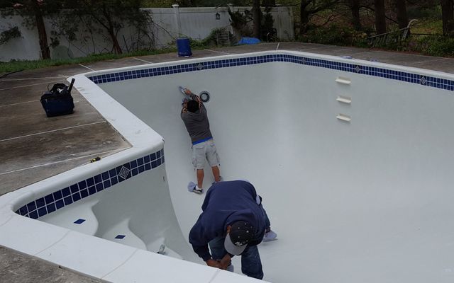 Two people working inside an empty pool with white walls and blue tile border.