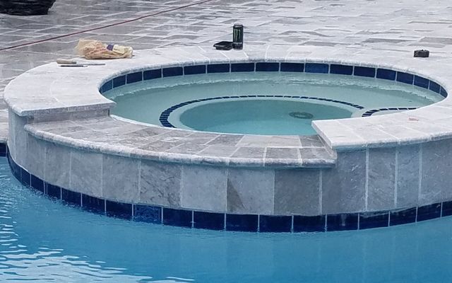 Hot tub with blue water, surrounded by light gray stone and dark blue tiles, next to a swimming pool.