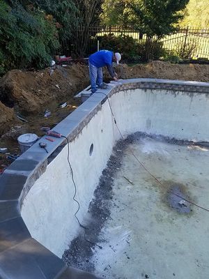Construction worker working on the edge of an empty pool. Concrete pool walls, gray coping, dirt and trees in background.