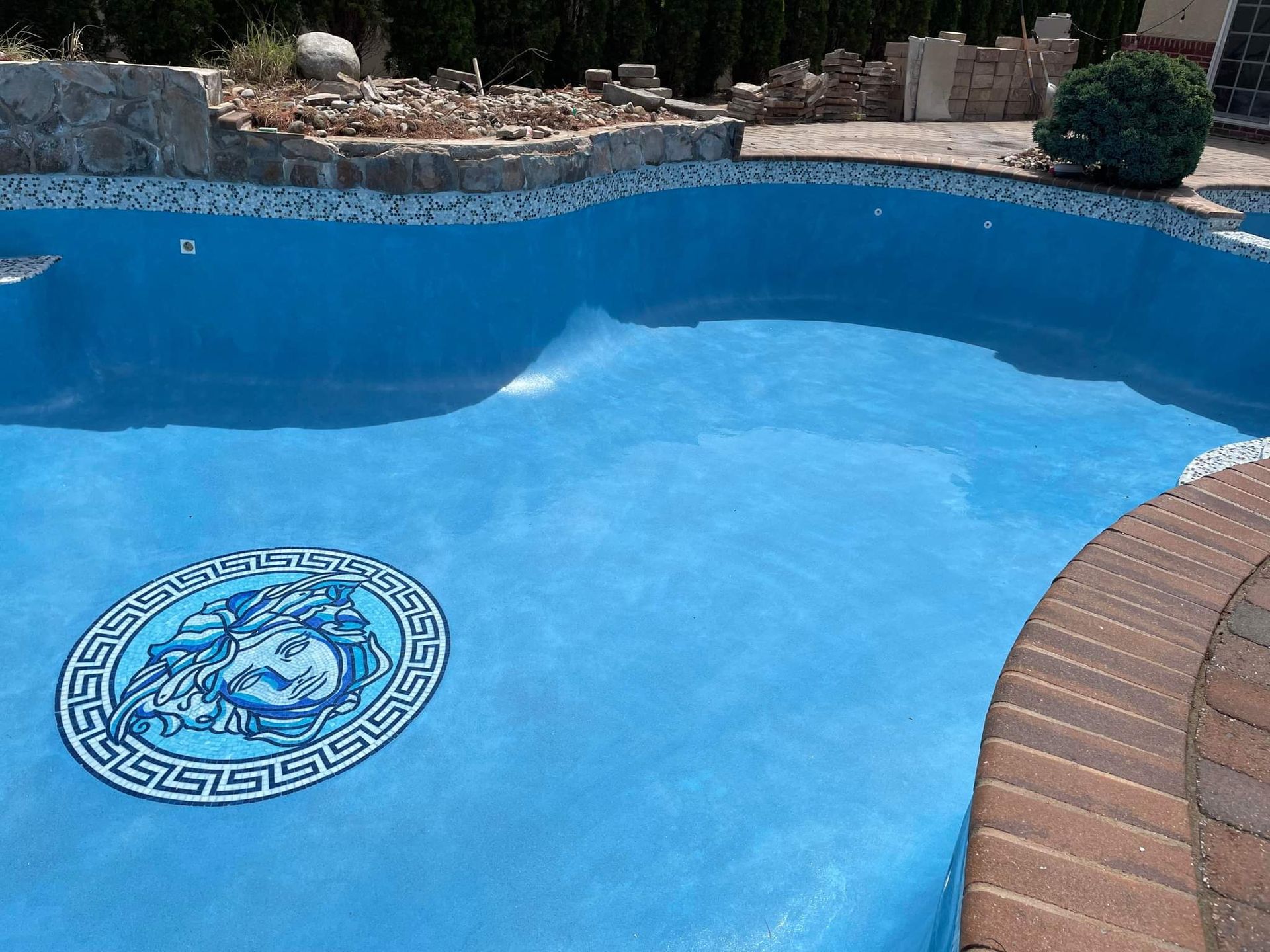 Blue swimming pool with mosaic Versace emblem. Brick coping and tile border visible.