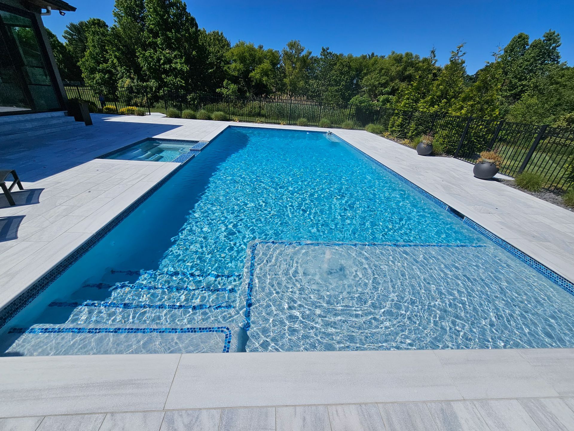 Rectangular swimming pool with light blue water and steps, surrounded by white stone patio and greenery under a clear sky.