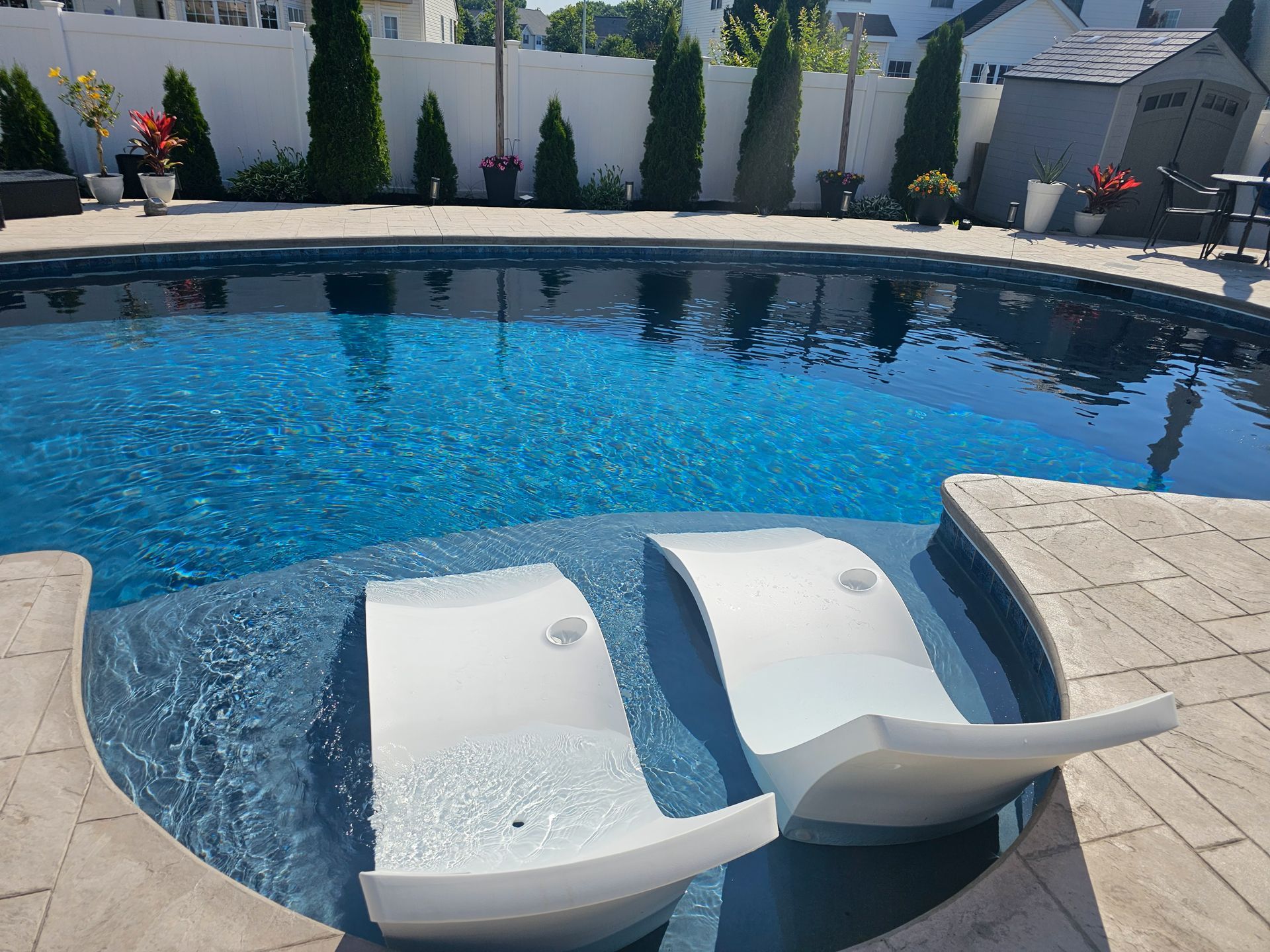 Two white lounge chairs in a blue swimming pool. Outdoor setting with a white fence and trees.