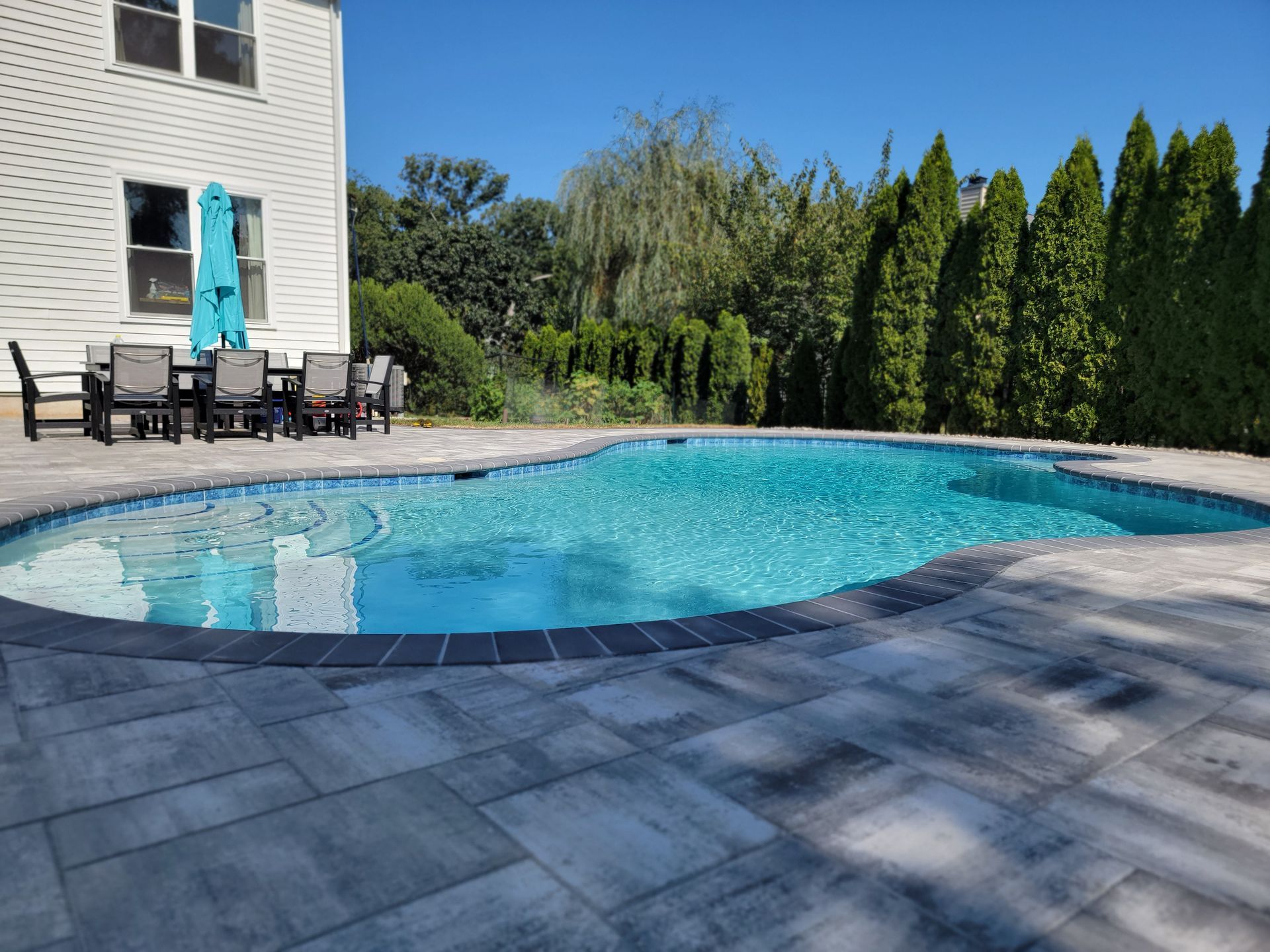 Swimming pool with patio furniture beside a two-story house, surrounded by pavers, trees, and blue sky.