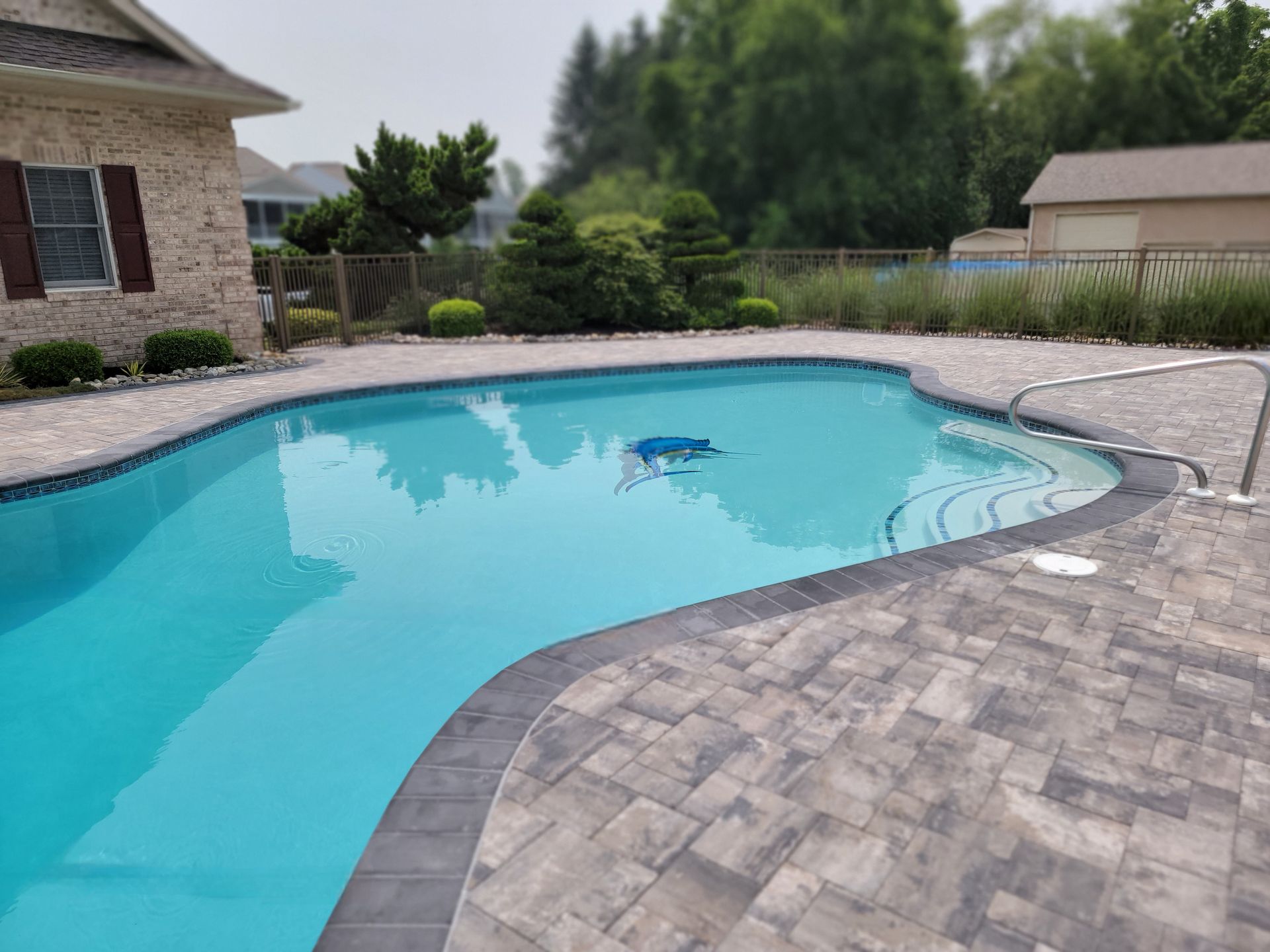 Pool with blue water surrounded by stone patio, landscaping, and a house.