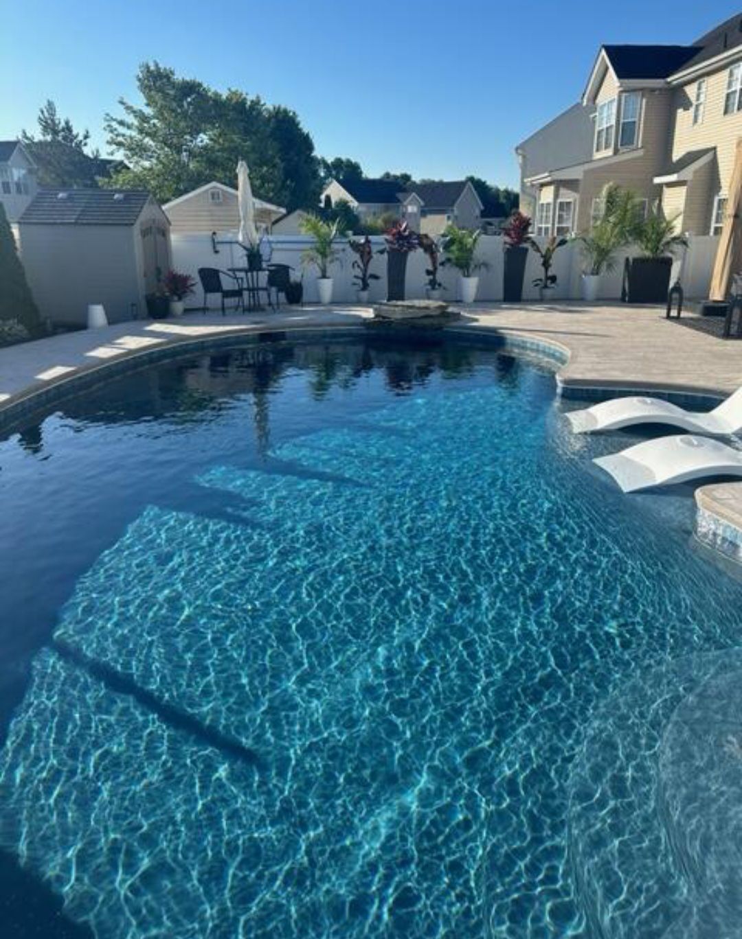 Swimming pool with clear blue water and lounge chairs, surrounded by patio and houses under a sunny sky.