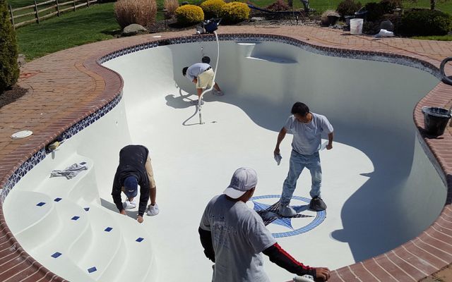 Four workers renovating an empty swimming pool. Pool has light-colored surfaces and a decorative emblem.