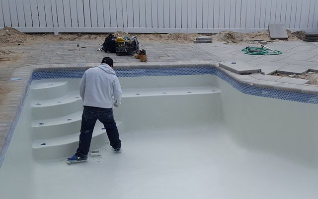 Person inside a newly painted swimming pool. Steps on the left, blue tile trim. White fence in the background.