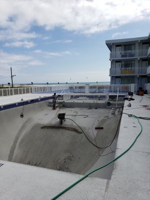 Pool under construction, next to a white building. Construction workers, tools, and a sea view visible.