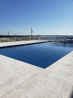 Outdoor pool with blue water and white concrete deck, blue sky, and ocean in the distance.
