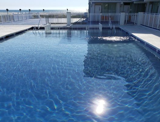 Blue swimming pool with sun reflection, white railing, and building.