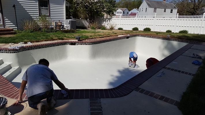 Men painting the inside of an empty swimming pool. Brick border, white walls, sunny outdoors.