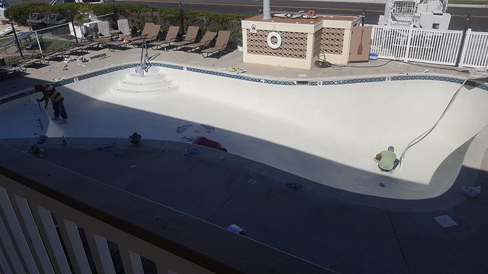 Empty swimming pool being worked on. Workers on the ground, beige plaster, white fence and building in the background.