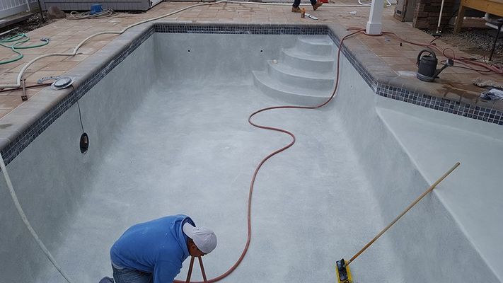 Workers resurfacing an empty swimming pool with white plaster. Blue tiled border and steps, hose present.